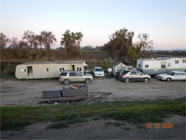 Photo taken through chainlink fence with Smith Canal in background.  Foreground shows cardboard box tent with blue tarps covering it. Levee rocks moved around encampment to protect from water. - Date of 03.11.2020 shows at the bottom of the photo