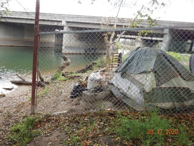 image of homeless tent and encampment on side of river behind chain link fence
