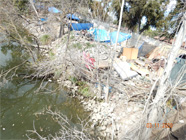 View of tarps, tents, and debris of homeless encampment along the Smith Canal edge shown through hole in chain link fence - Date of 03.11.2020 shows at the bottom of the photo