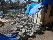 Photo taken through chainlink fence with Smith Canal in background.  Foreground shows cardboard box tent with blue tarps covering it. Levee rocks moved around encampment to protect from water. - Date of 03.11.2020 shows at the bottom of the photo