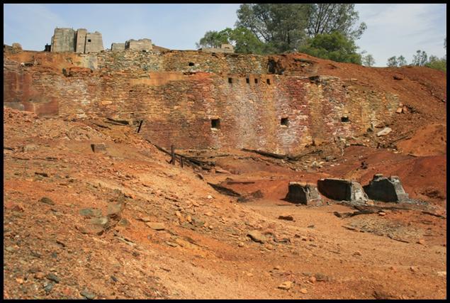 small hill and cliff made of red dirt.  Rock structure with small square holes in side of cliff.  Tree on top of cliff. Blue sky in background.