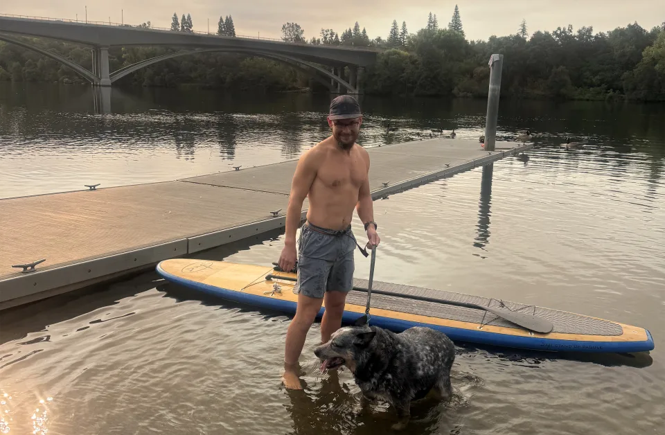 A paddleboarder standing in shallow water beside his paddleboard and his dog at Black Miners Bar